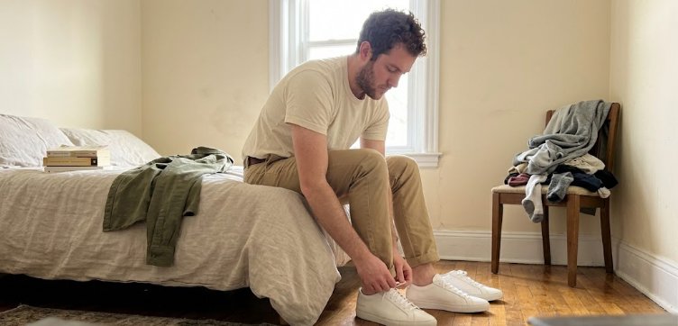 Man putting on white sneakers wearing cream tee and khaki chinos at home
